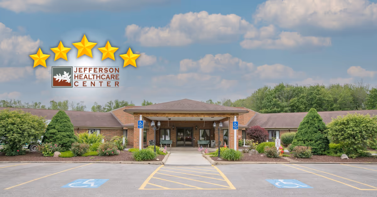Front exterior view of Jefferson Healthcare Center, a single-story brick building with a covered entrance, surrounded by landscaped bushes and trees under a partly cloudy sky. There are two handicap parking spaces visible in the foreground.