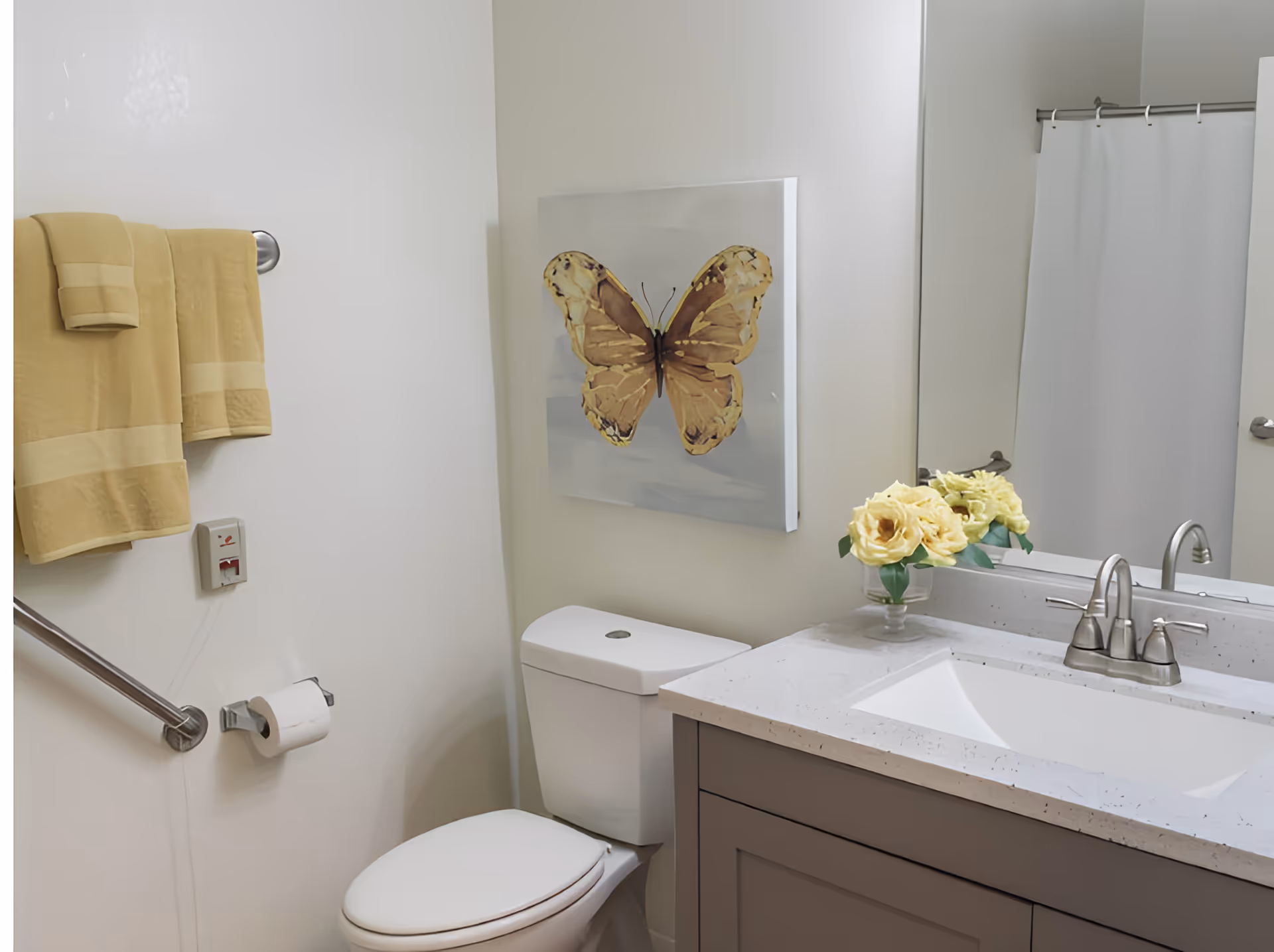 A bright bathroom with a toilet, vanity sink and mirror, yellow towels, butterfly wall art, and a vase of yellow flowers.