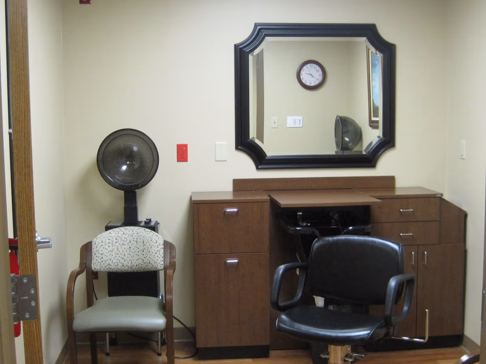 Interior of a small salon room with a black salon chair in front of a wooden counter with drawers and a large decorative mirror above it. To the left, there is a beige chair with wooden armrests and a vintage hair dryer behind it. The walls are light-colored, and a clock is visible in the mirror's reflection.