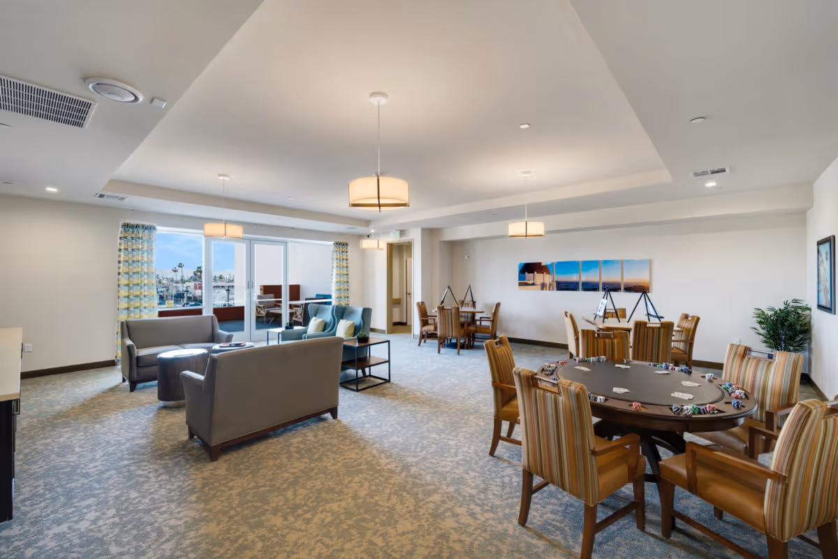 A spacious senior living common area with comfortable seating including sofas and armchairs, a poker table with chairs and poker chips, and additional tables and chairs in the background. The room has large windows with patterned curtains allowing natural light, modern ceiling lights, and artwork on the walls.