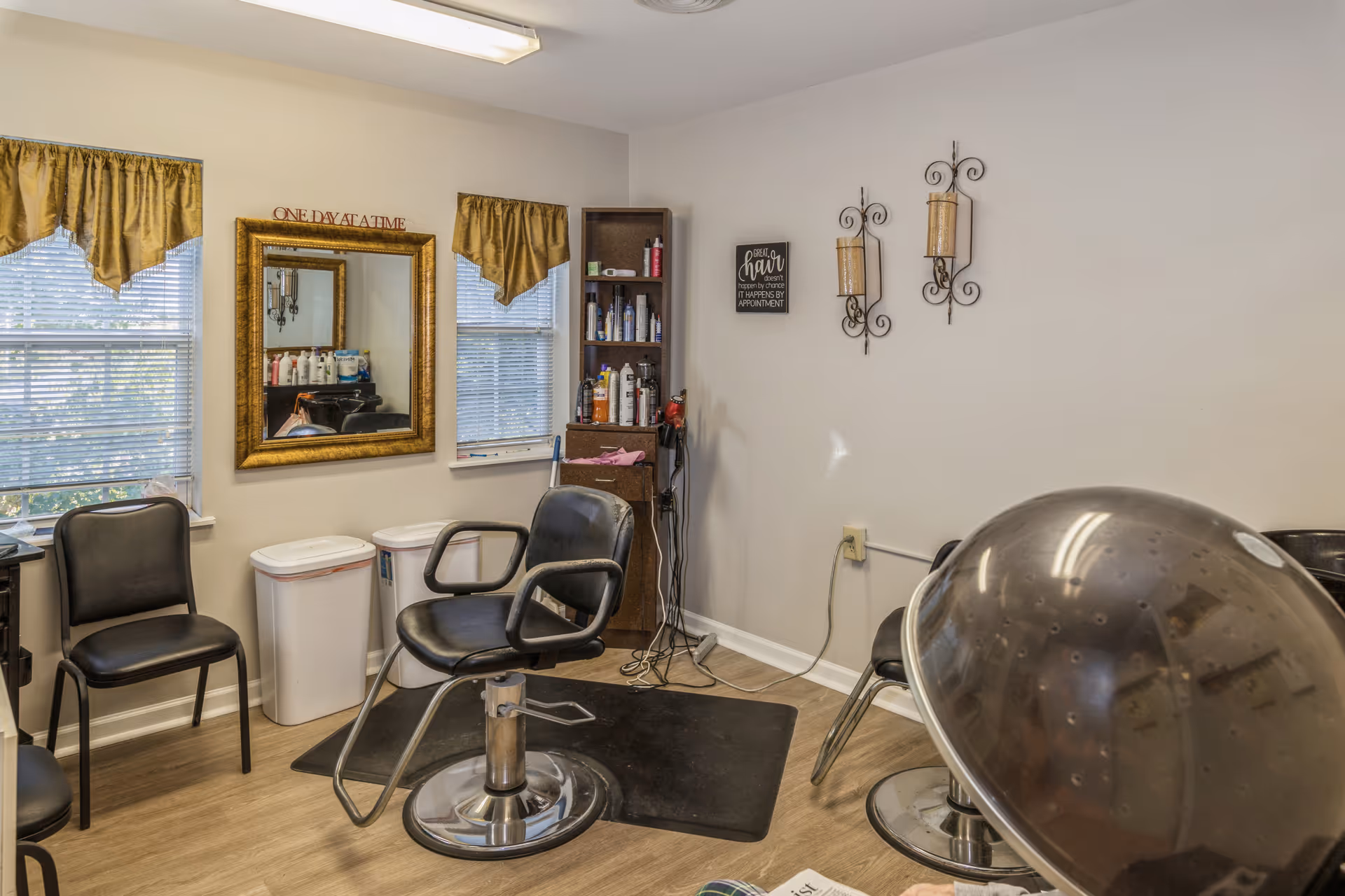 Interior of a small hair salon with two black salon chairs, a large mirror with a gold frame on the wall, two windows with gold curtains, shelves with hair products, and a hair dryer hood in the foreground. The walls are decorated with candle holders and a small sign.