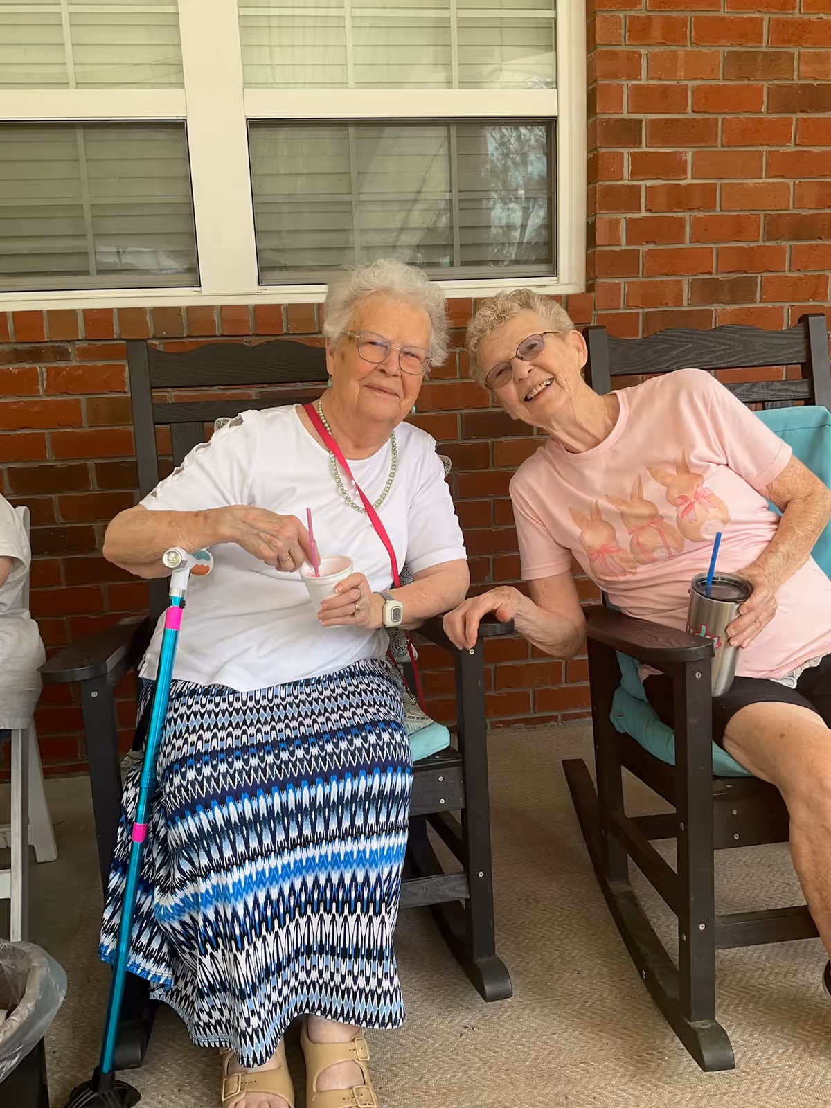 Two elderly women sitting on wooden rocking chairs on a porch with a brick wall and window behind them. One woman is wearing a white shirt and patterned skirt, holding a cup and spoon, with a blue cane resting beside her. The other woman is wearing a pink shirt with a bunny design and black shorts, holding a tumbler with a straw. Both women are smiling and appear to be enjoying each other's company.