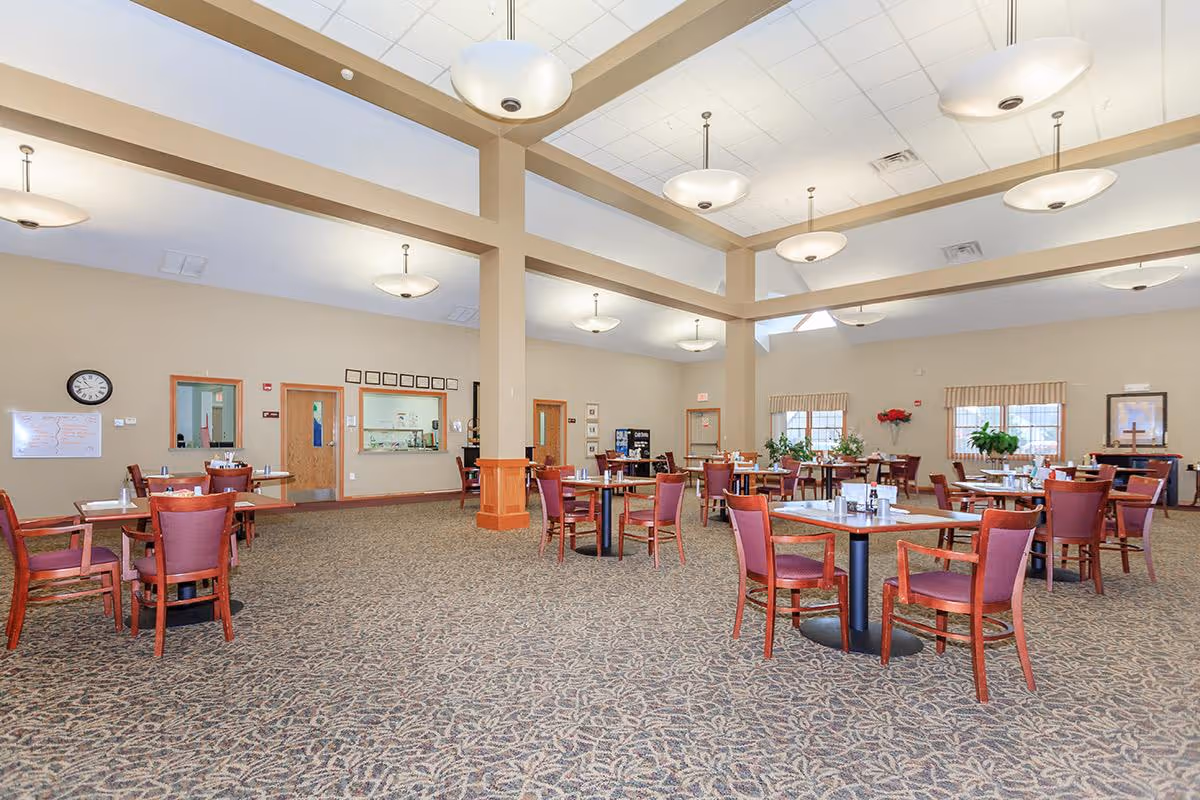 A spacious dining room with multiple round tables and wooden chairs with red cushions. The room has a high ceiling with several hanging light fixtures, beige walls, and patterned carpet. There are windows with valances, plants, and a clock on the wall. A serving window and doors are visible in the background.