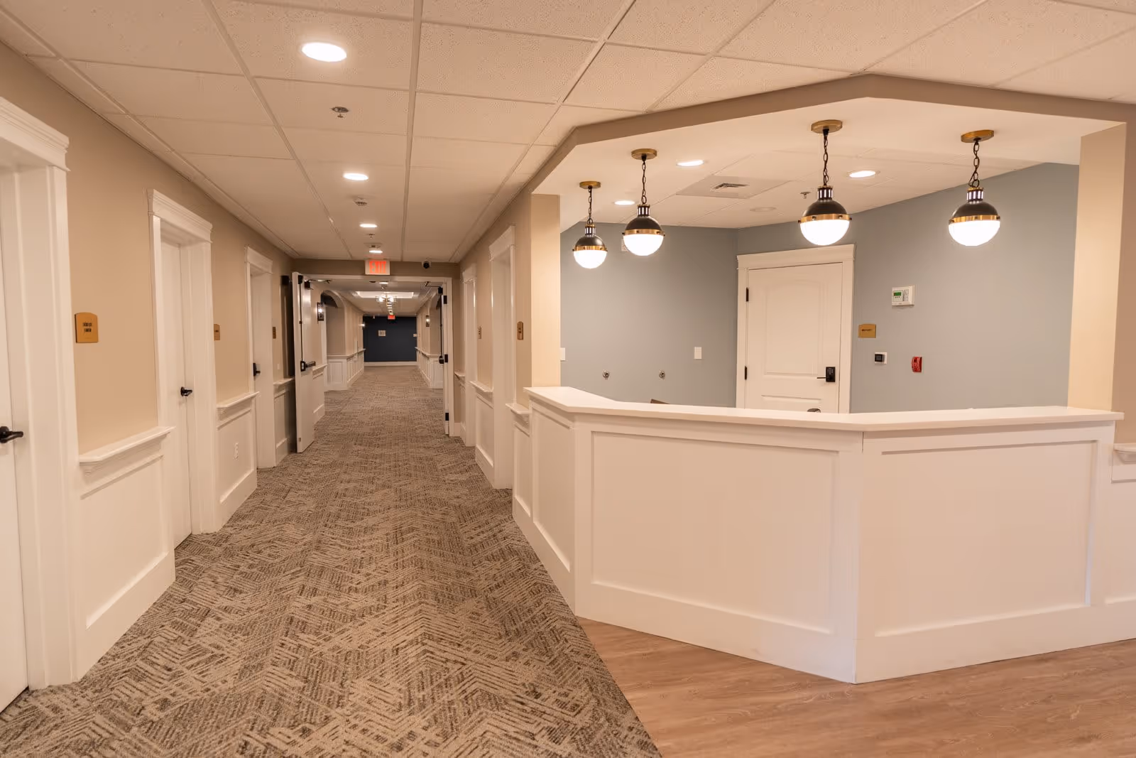 Interior hallway of a senior living facility with beige walls, patterned carpet, and white doors on both sides. On the right side, there is a white reception desk area with three hanging pendant lights and a door behind it. The ceiling has recessed lighting and an exit sign is visible at the far end of the hallway.