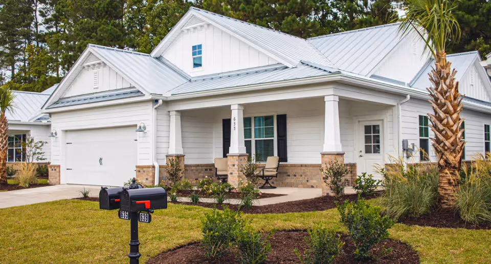 White single-story house with a metal roof, front porch with chairs, attached garage, and mailboxes by the lawn.