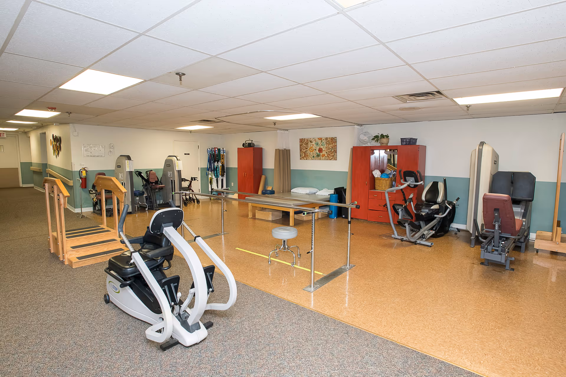 A senior living facility physical therapy/exercise room with exercise machines, parallel bars, ramps, and storage cabinets.
