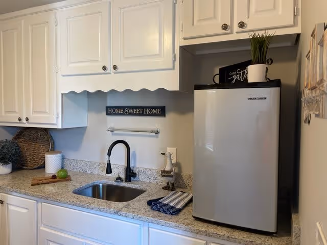 A small kitchen area with white cabinets and granite countertops. There is a stainless steel sink with a black faucet, a mini refrigerator on the right side, and decorative items including a 'HOME SWEET HOME' sign on the wall, a small plant on top of the refrigerator, and a cutting board with fruit on the counter.
