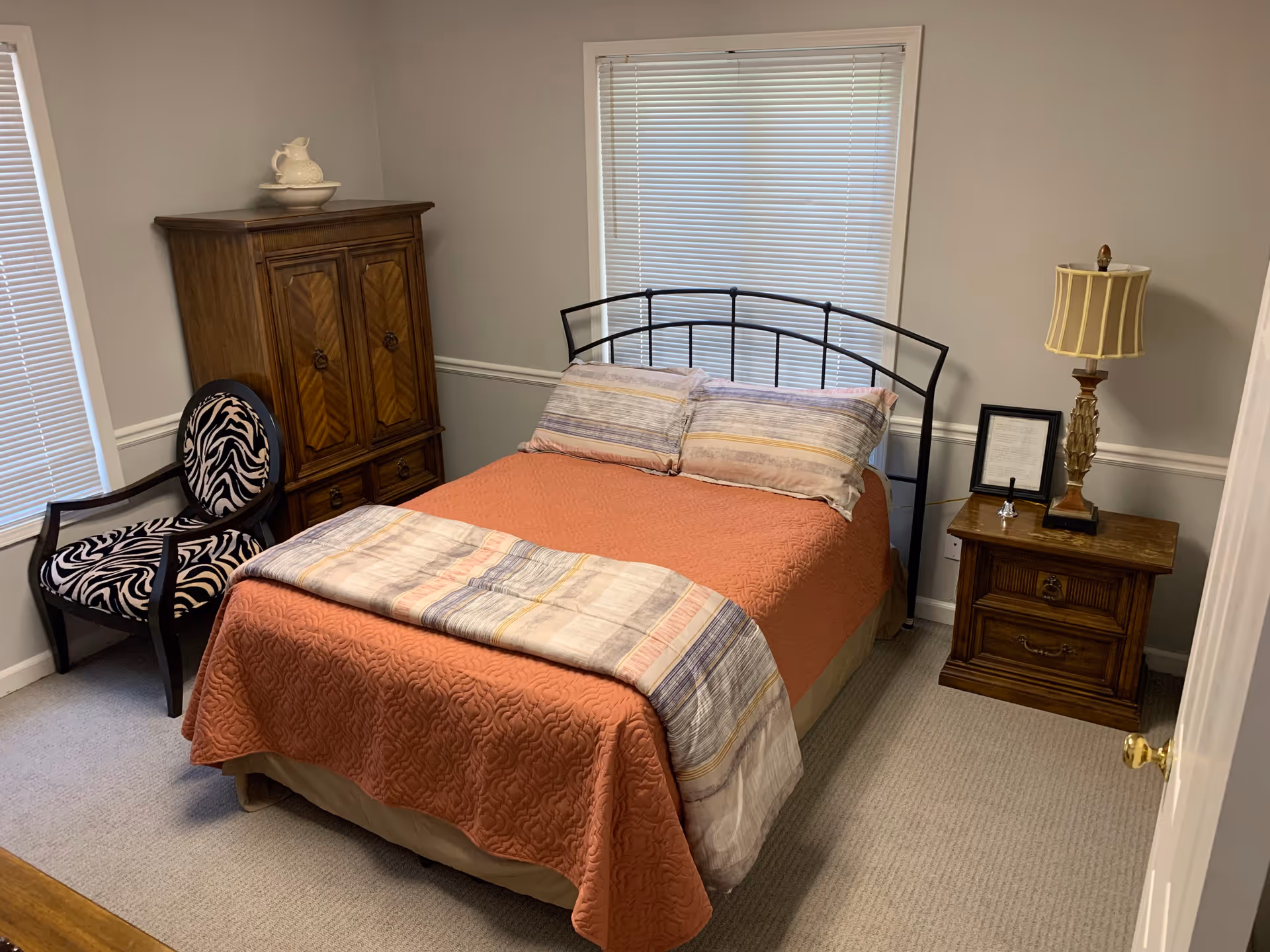 A bedroom with a bed covered in an orange quilt and striped pillows and blanket. There is a wooden nightstand with a lamp and framed picture on the right side of the bed. To the left, there is a wooden armoire and a black chair with zebra-patterned upholstery. The room has light gray walls, two windows with blinds, and beige carpeted floor.