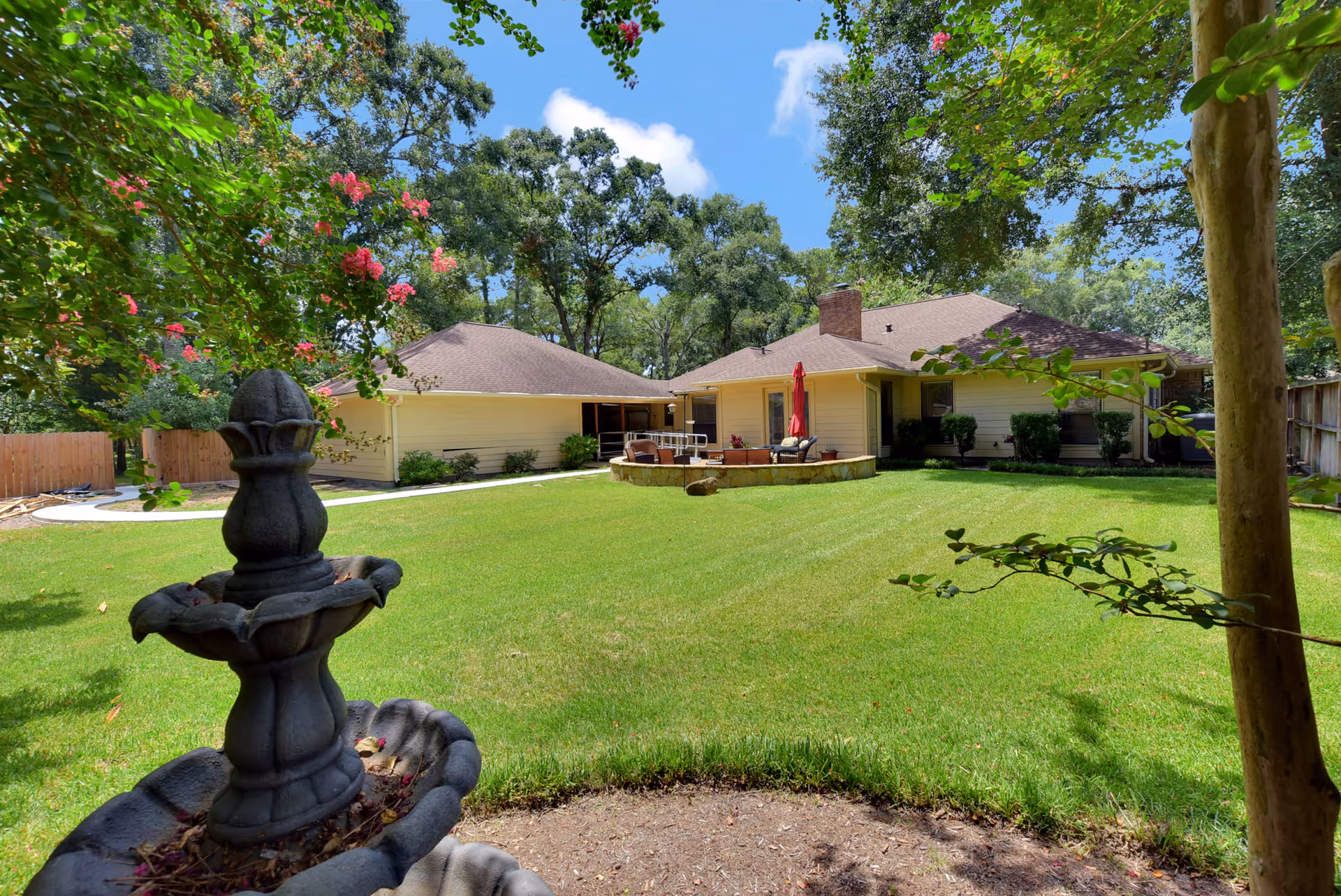 Well-kept backyard with a stone fountain in the foreground, a large grassy lawn, and a single-story house with a patio and umbrella in the background surrounded by trees.