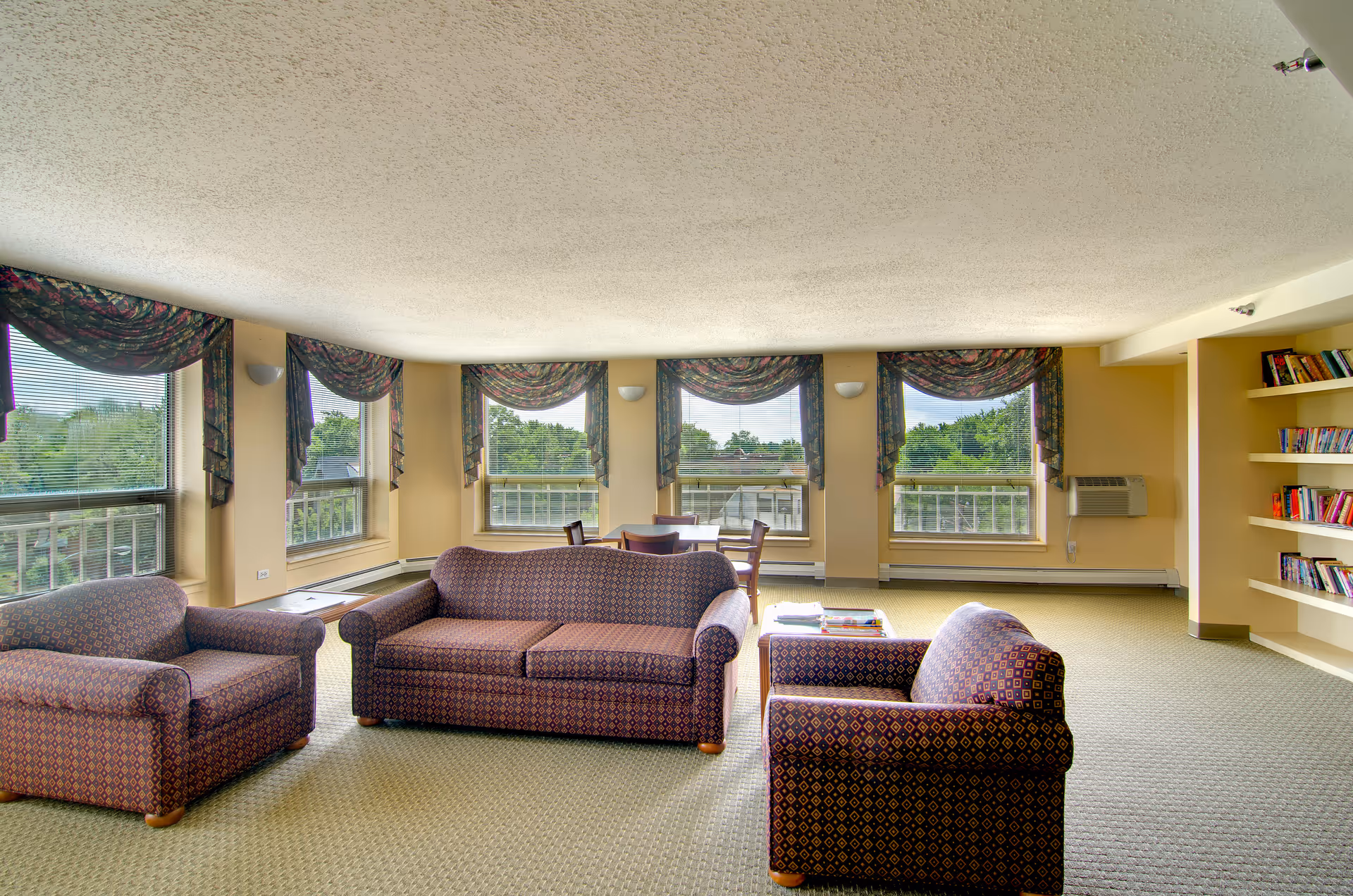 A spacious living room area with three patterned upholstered chairs and a sofa arranged around a small table with books. The room has large windows with floral valances, allowing natural light to fill the space. There is a small dining table with chairs near the windows and built-in bookshelves on the right wall filled with books. The walls are painted a light beige color and the floor is carpeted.