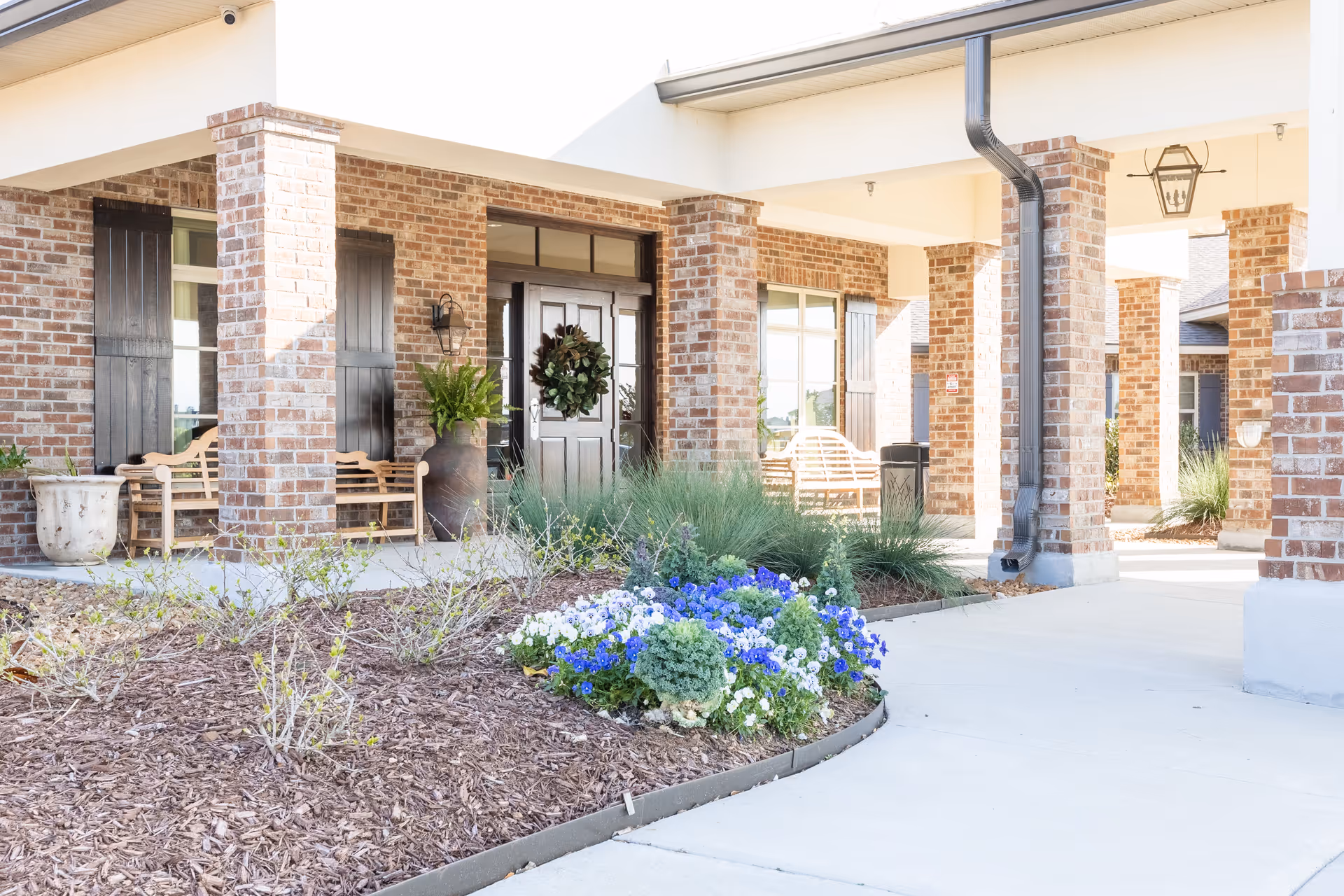 Entrance of a senior living facility with brick pillars and walls, black double doors decorated with a wreath, wooden benches on the porch, potted plants, and a flower bed with blue and white flowers in the foreground.