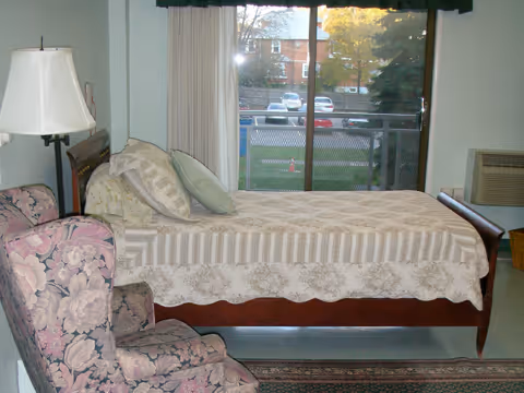 A bedroom with a single bed covered with a patterned bedspread and pillows. Next to the bed is a floral upholstered armchair and a floor lamp. A large window with curtains shows a view of a parking lot and trees outside.