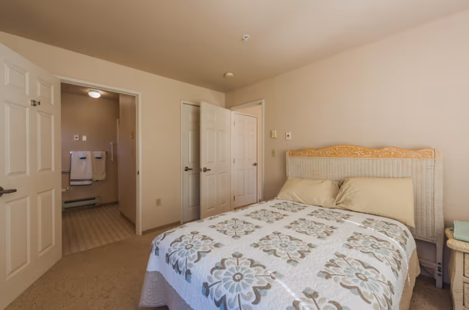 A simple bedroom with a floral quilted bed, wicker headboard, and open doors leading to a bathroom and hallway.