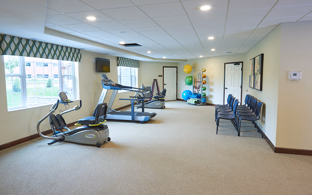 A bright exercise room with large windows, exercise equipment including a recumbent bike, treadmill, and elliptical machine, colorful exercise balls and weights on racks, and a row of blue chairs along the wall.