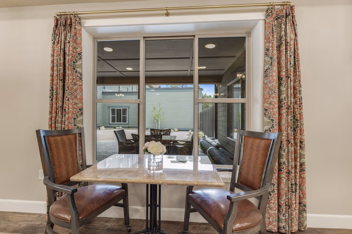 A small dining area with a square marble table and two wooden chairs with brown leather seats and patterned backs. The table has a small glass vase with white and pink flowers. Behind the table is a large window with patterned curtains, showing an outdoor patio area with additional seating and a building exterior.