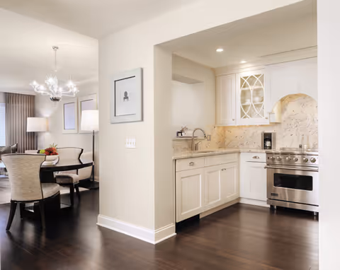 View of a modern kitchen with white cabinets, marble backsplash, stainless steel stove, and a sink. Adjacent to the kitchen is a dining area with a round table, upholstered chairs, a chandelier, and a floor lamp. The flooring is dark hardwood throughout.