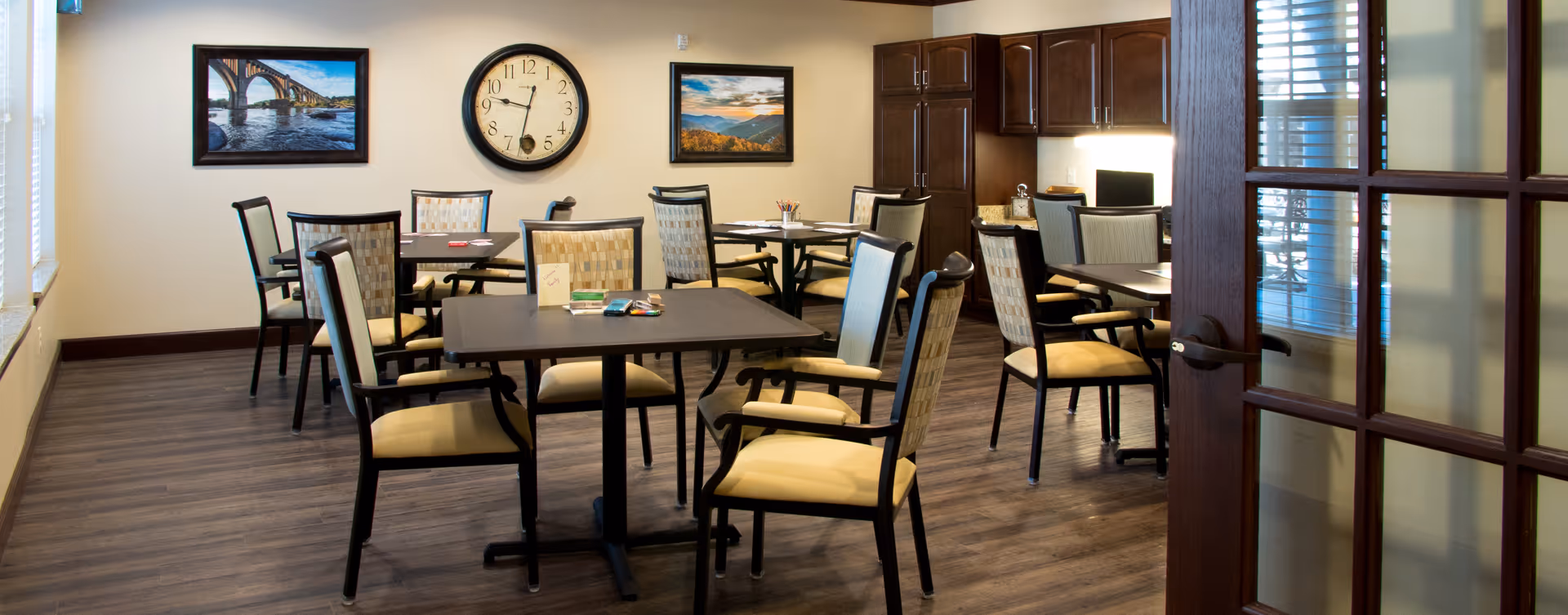 A well-lit room with several tables and cushioned chairs arranged neatly on a wooden floor. The walls are decorated with two framed landscape photographs and a large round clock. There are dark wooden cabinets and a desk with a computer in the corner. The room is viewed through a partially open wooden door with glass panels.
