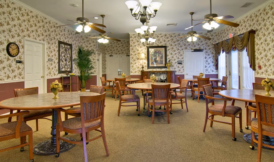 A senior living facility dining room with multiple round tables and wooden chairs arranged neatly. The room features floral wallpaper, ceiling fans with lights, chandeliers, a clock on the wall, framed artwork, and large windows with curtains allowing natural light to enter.