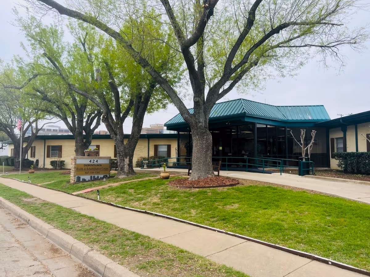 Exterior view of Downtown Health and Rehabilitation Center showing a single-story building with a green roof, large trees, a sidewalk, and a sign with the facility's name and phone number.