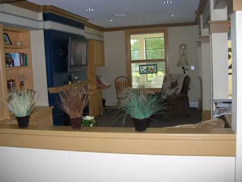 Interior view of a senior living lounge with seating, plants, and a window.