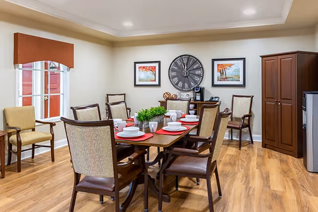 A dining room with a wooden table set for six people with white plates, bowls, and cups on red placemats. The room has wooden flooring, beige walls, and a large clock on the wall flanked by two framed pictures of autumn trees. There are several upholstered chairs around the table and along the walls, a wooden cabinet, and a small refrigerator.