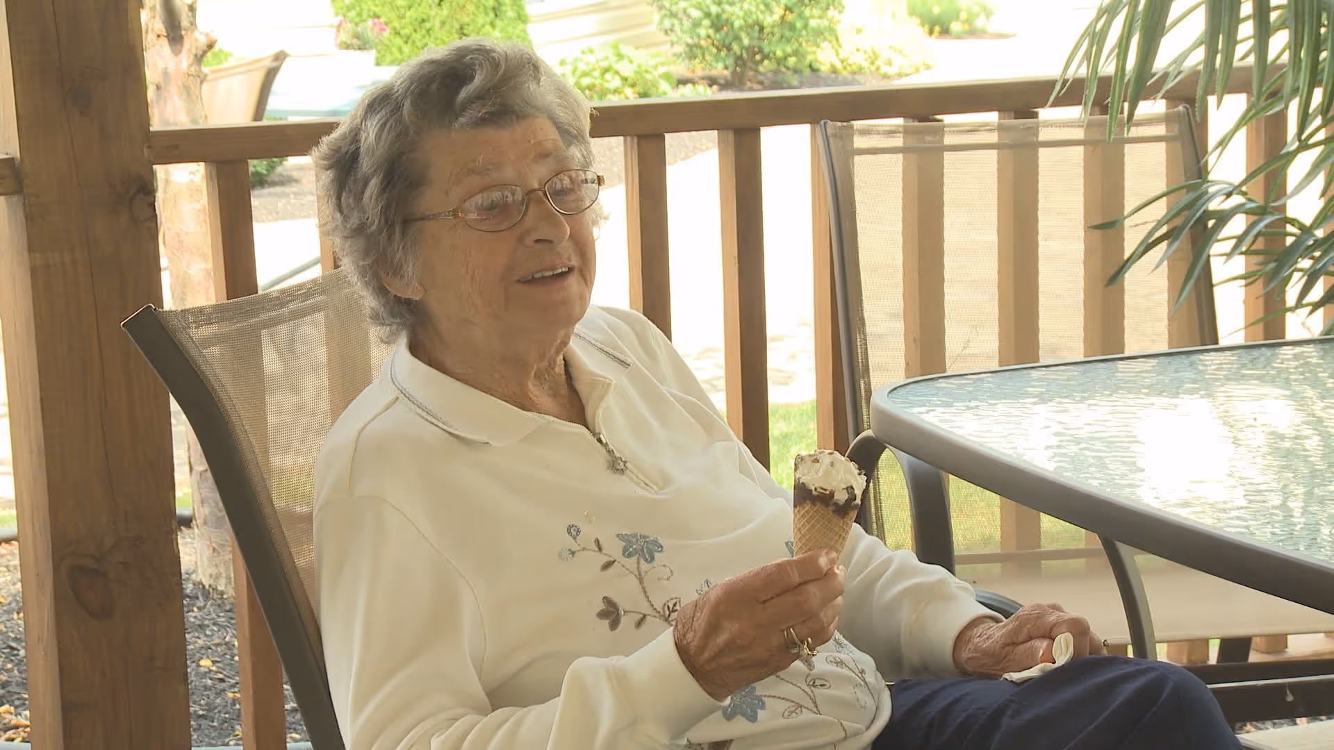 An elderly woman wearing glasses and a white sweater with floral embroidery is sitting on a mesh chair outdoors under a wooden pergola, holding an ice cream cone with chocolate and whipped cream. There is a glass-top table and another chair nearby, with greenery and a garden visible in the background.