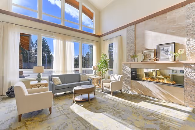 A bright and spacious living room with large windows letting in natural light. The room features a gray sofa, two beige armchairs, a round coffee table, two side tables with lamps, a modern fireplace with decorative items on the mantel, and a potted plant near the window. The walls are light-colored and the carpet has a subtle pattern.