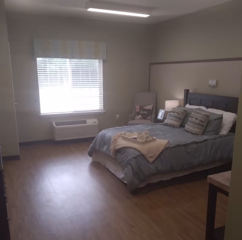 A cozy bedroom with a neatly made bed featuring blue bedding and decorative pillows. There is a tray with cups and a blanket on the bed. Next to the bed is a nightstand with a lamp and a framed photo. A cushioned armchair with a decorative pillow is placed near a window with blinds and a valance. The room has wood flooring and neutral-colored walls.
