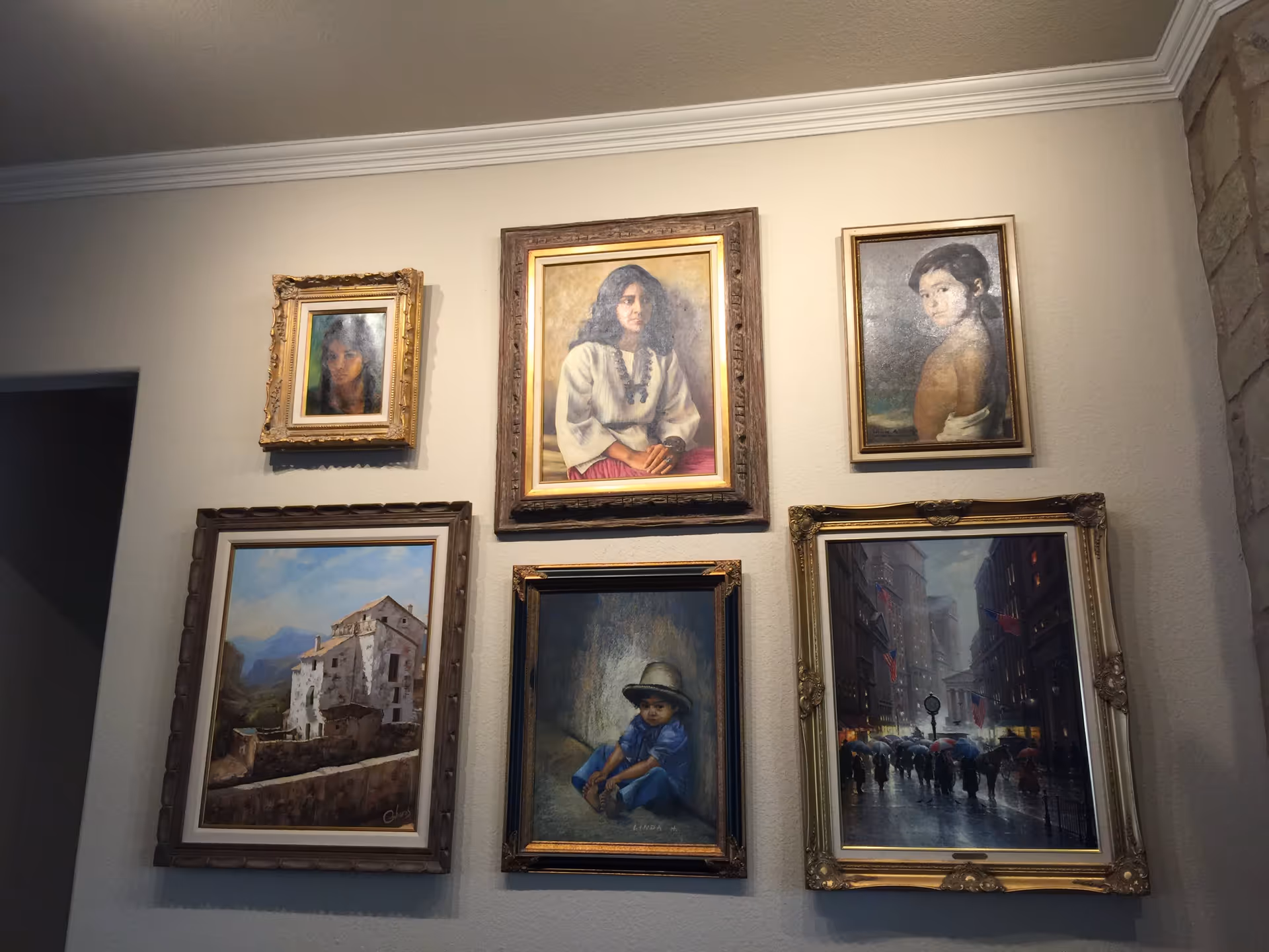A wall displaying six framed paintings, including portraits of people and scenes of buildings and a city street with people holding umbrellas. The wall has a beige color and a crown molding at the ceiling. A stone wall is partially visible on the right side.