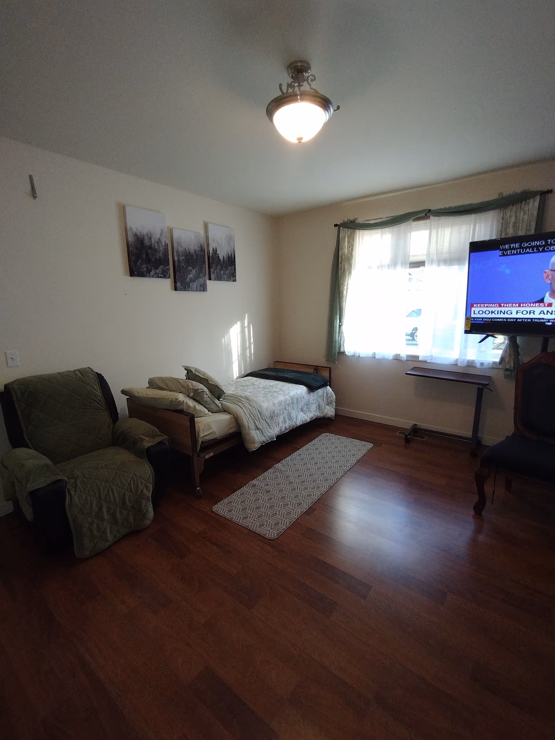 A small bedroom with a single bed covered with a light-colored blanket and pillows, a green armchair with a matching cover, a small rug on the wooden floor, a window with sheer curtains letting in natural light, a wall-mounted TV showing a news program, and three black and white landscape pictures on the wall.
