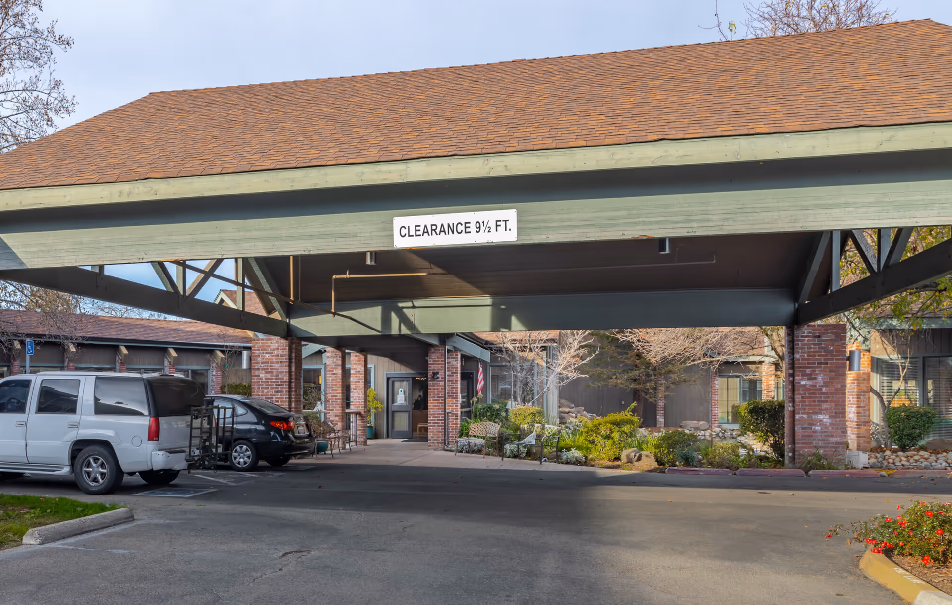 Covered driveway entrance to a building with a clearance sign of 9 1/2 feet. There are parked cars on the left side and benches with plants on the right side near the entrance. The building has brick pillars and a brown shingled roof.