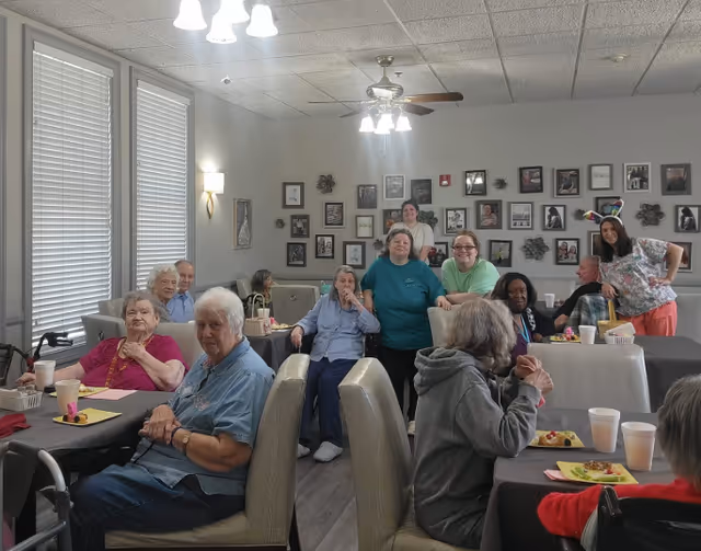 Seniors and caregivers seated and standing around tables in a bright communal dining room with framed photos on the wall.