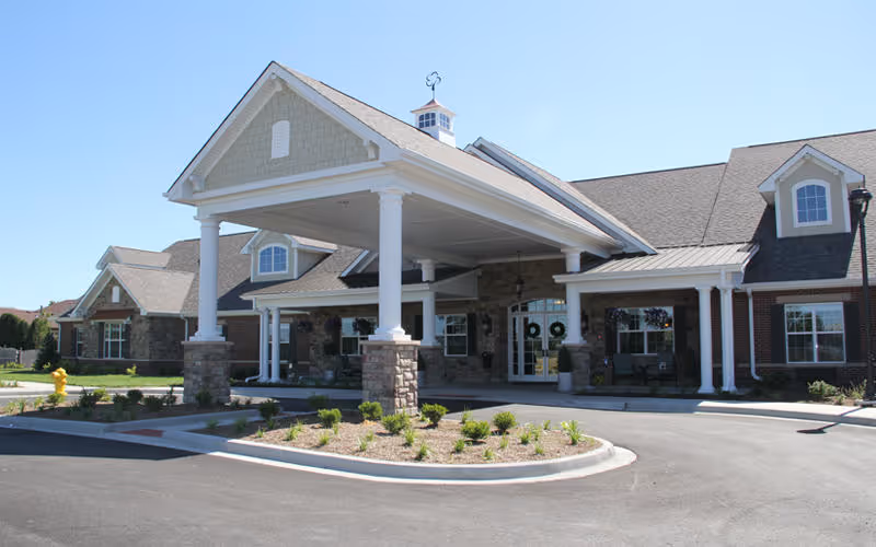 Exterior view of a senior living facility building with a large covered entrance supported by white columns and stone bases. The building has a mix of brick and stone facade with multiple windows and a well-maintained landscaped area in front. The sky is clear and blue.