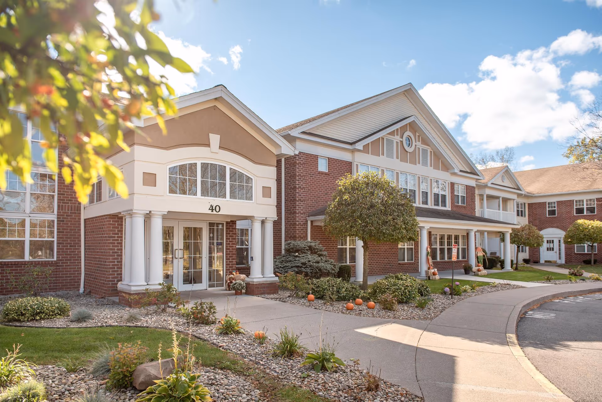 Exterior view of a senior living facility building with red brick walls, white columns, and large windows under a blue sky with some clouds. The entrance has the number 40 above the door, and there are small trees, shrubs, and pumpkins decorating the landscaped area around the walkway.