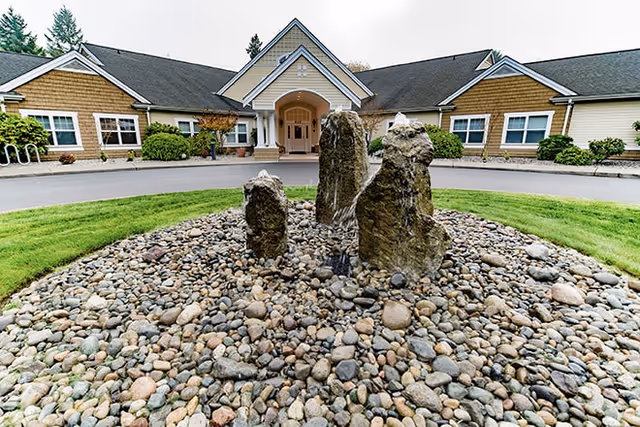 Front exterior view of a single-story building with a peaked roof and a covered entrance. In the foreground, there is a circular rock garden with several large standing stones surrounded by smaller pebbles. The building is surrounded by green grass and some shrubs.