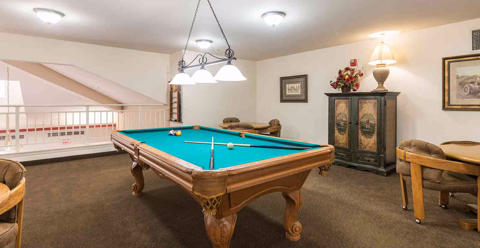 A recreational room with a green felt pool table in the center, two pool cues and a white cue ball on the table. The room has beige walls, carpeted floor, and ceiling lights. There are two round tables with cushioned chairs around them, a decorative cabinet with a lamp and flower arrangement on top, and framed artwork on the walls.