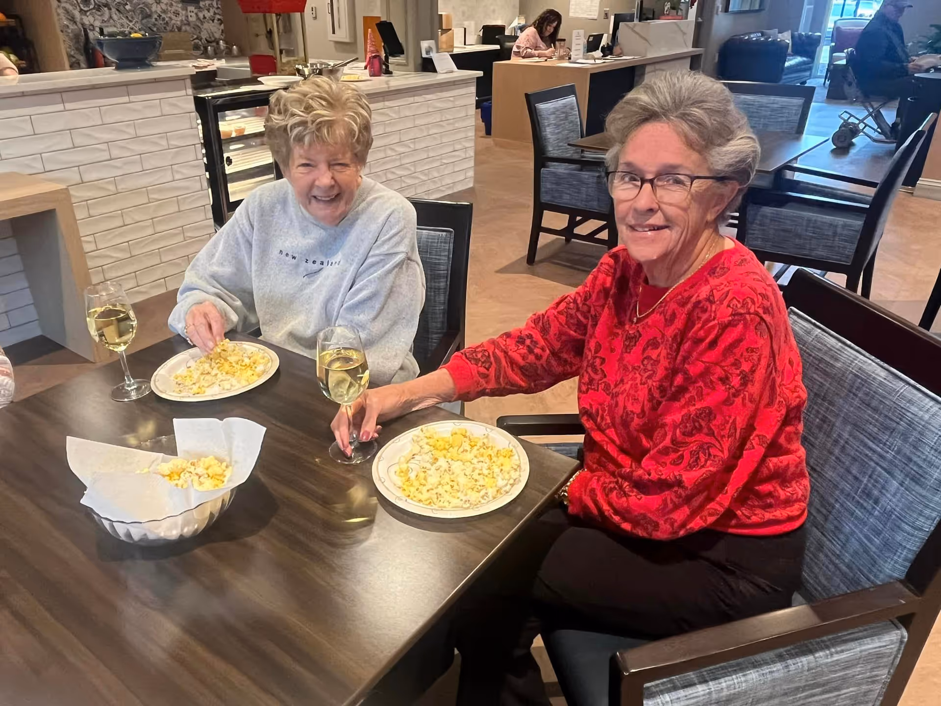 Two elderly women sitting at a table in a dining area, each with a plate of popcorn and a glass of white wine. One woman is wearing a gray sweatshirt and smiling broadly, while the other is wearing a red patterned sweater and glasses, smiling at the camera. In the background, there are other tables, chairs, and people, including a woman working at a desk.