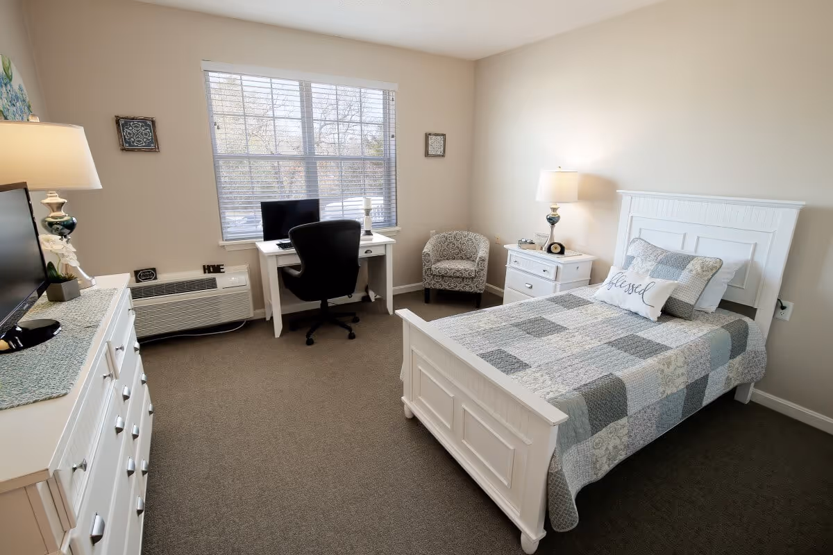 A bright and tidy bedroom featuring a single bed with a patchwork quilt and a pillow that says 'blessed'. The room includes a white dresser with a lamp and TV on top, a desk with a computer and office chair near a window with blinds, a patterned armchair in the corner, and a nightstand with a lamp and clock.