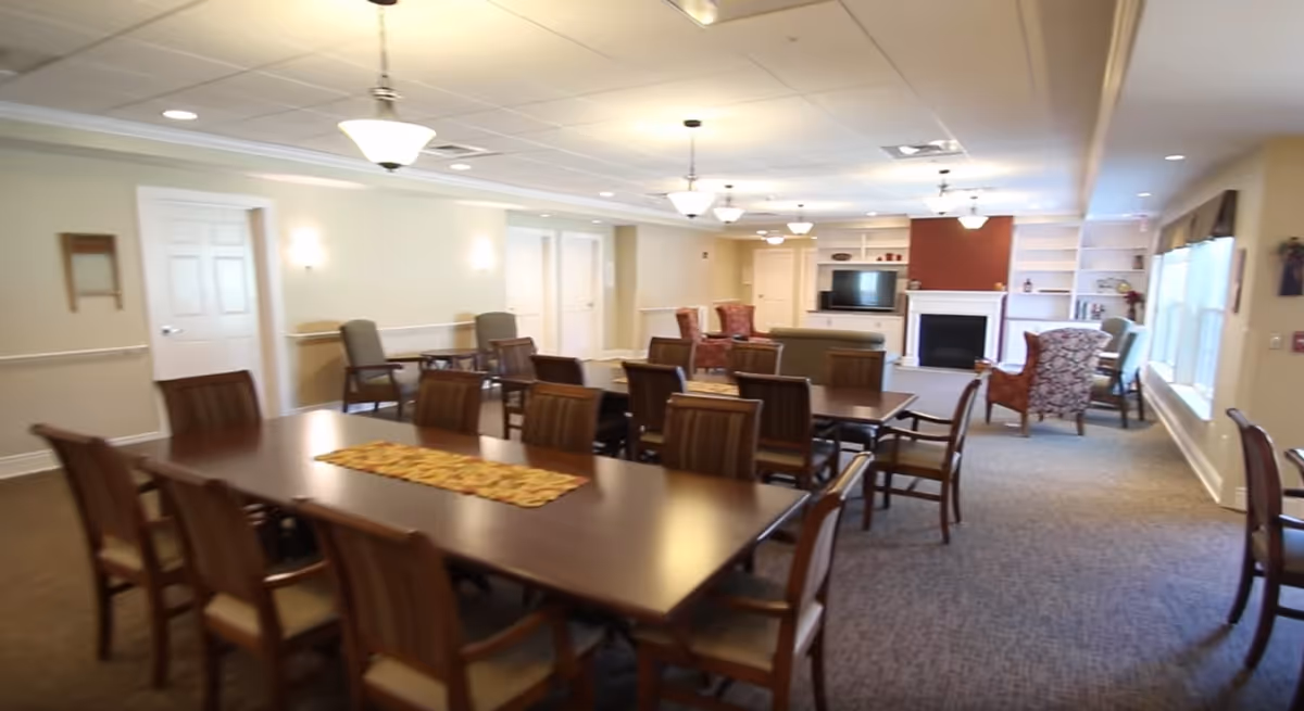 Spacious senior living common room with long dining tables and chairs leading to a seating area with a fireplace and television.