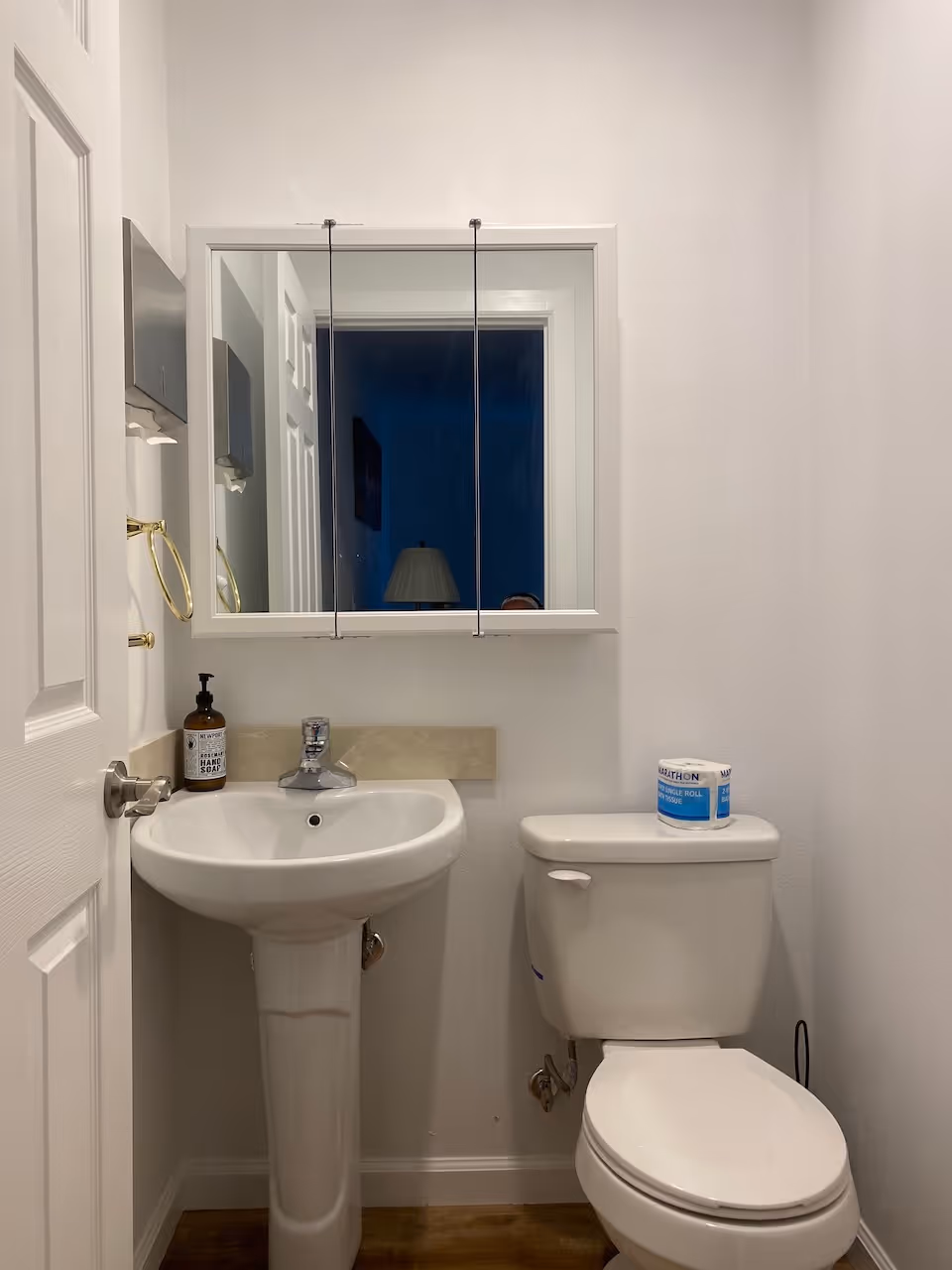 A small bathroom with a white pedestal sink and a white toilet. Above the sink is a mirrored medicine cabinet. There is a bottle of hand soap on the sink and a roll of toilet paper on top of the toilet tank. The walls are white and the floor is wooden.