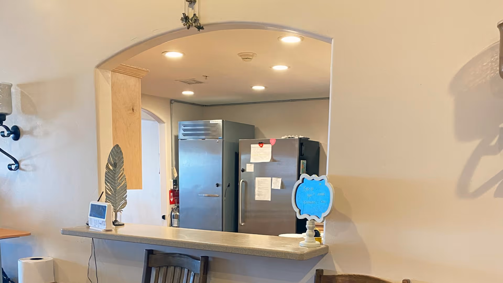 Interior view of a communal kitchen pass-through with a countertop, stools, and stainless steel refrigerator in the background.