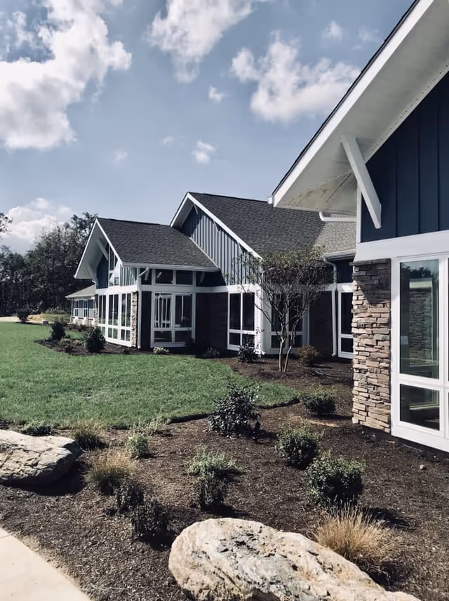 Exterior view of a modern senior living facility building with blue siding, white trim, large windows, stone accents, landscaped garden beds with small shrubs, and a green lawn under a partly cloudy sky.