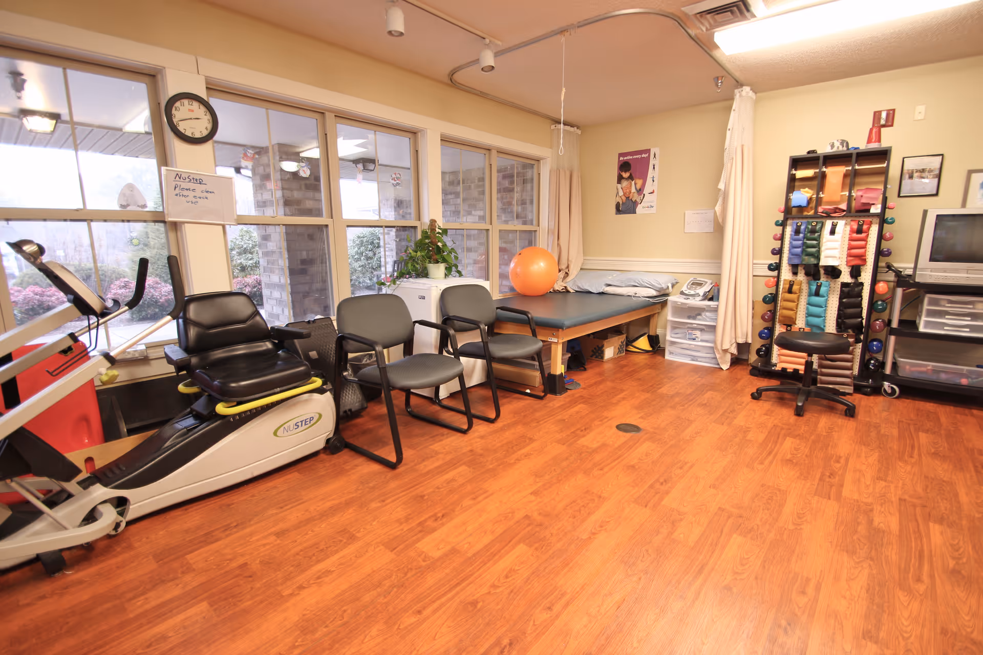 Therapy/exercise room with a NuStep recumbent stepper, a row of chairs, a treatment table, an exercise ball, and a rack of colorful hand weights.