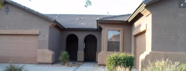 Exterior view of a single-story residential building with a garage on the left, an arched entryway in the center, and a window with a small bush in front on the right side.