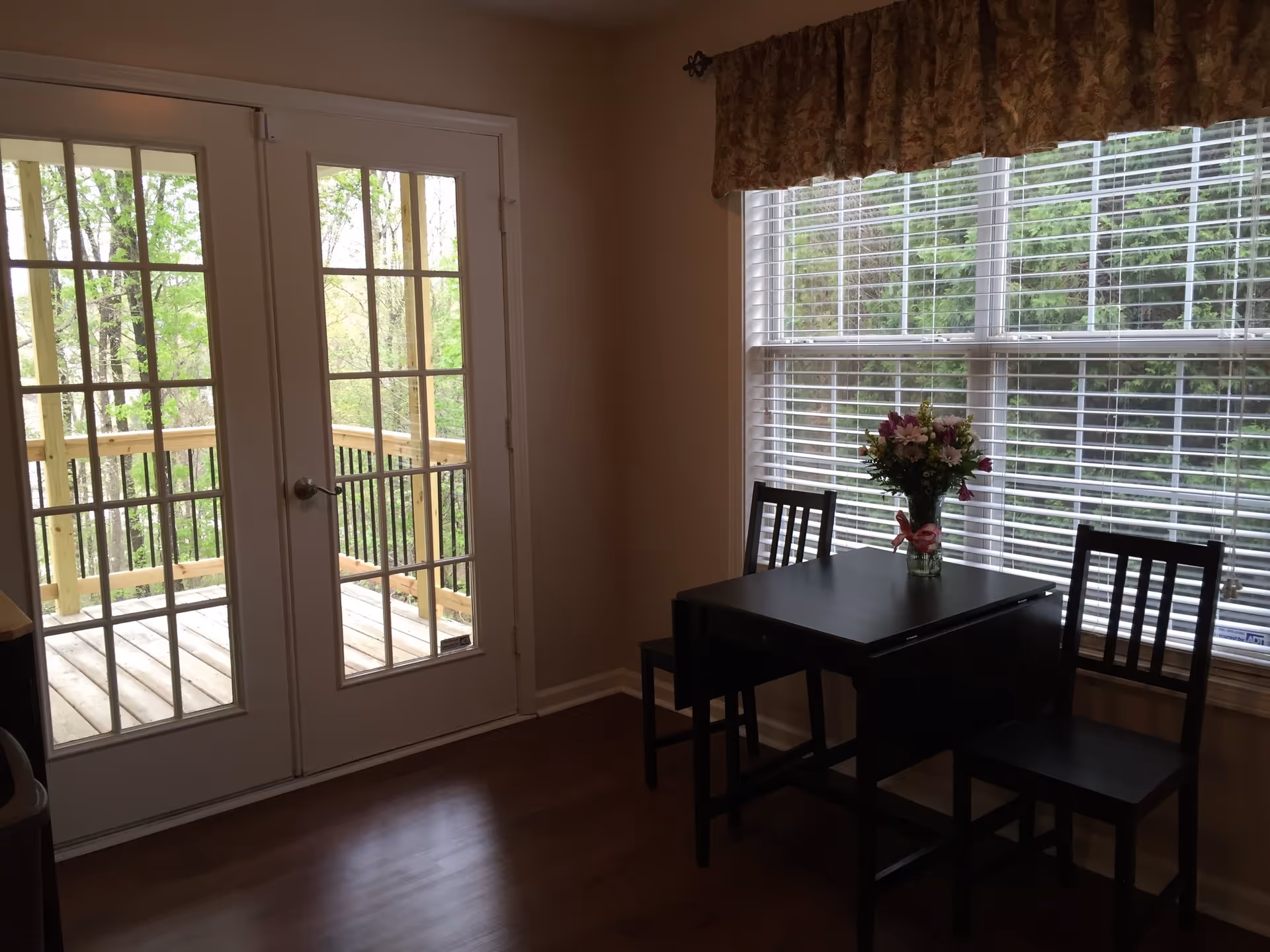 Small dining area with a black table and two black chairs next to a large window with white blinds. A vase with flowers is placed on the table. French doors lead to a wooden deck outside with green trees visible.