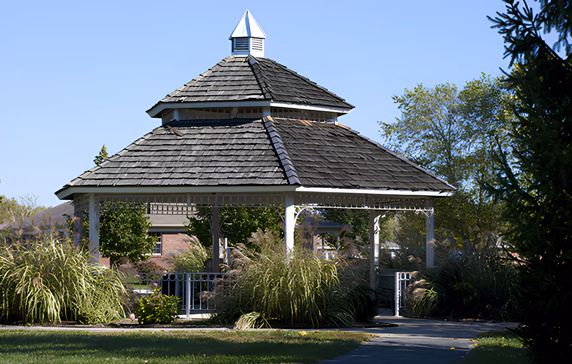 A white wooden gazebo with a multi-tiered shingled roof surrounded by ornamental grasses and trees on a sunny day.
