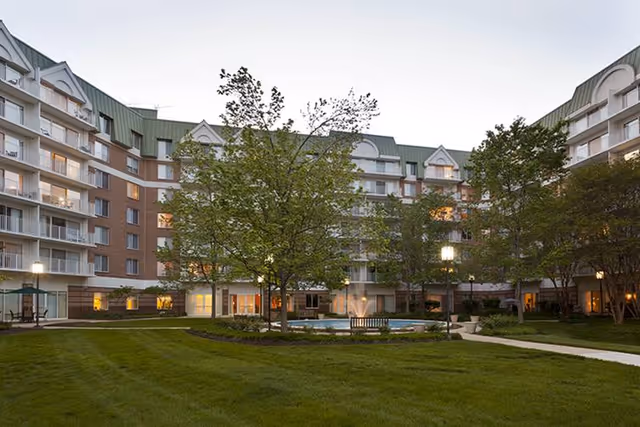 Outdoor courtyard area of a senior living facility named Bedford Court, featuring a well-maintained lawn, trees, benches, and a multi-story building surrounding the courtyard with balconies and lit windows at dusk.