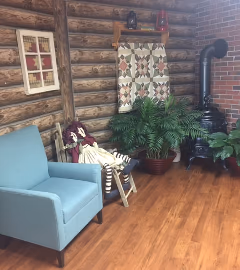 Cozy interior corner with a blue armchair, a rag doll on a small chair, potted plants, a wood stove, and log-cabin style walls with a hanging quilt.