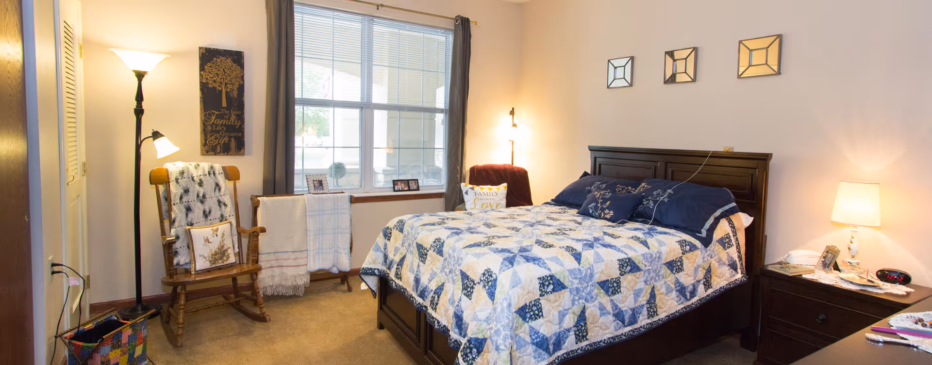 A cozy bedroom with a large window, a bed with a blue and white patterned quilt, a wooden rocking chair with a blanket, a floor lamp, and a nightstand with a lamp and alarm clock. The walls are decorated with small square mirrors and a family-themed wall hanging.