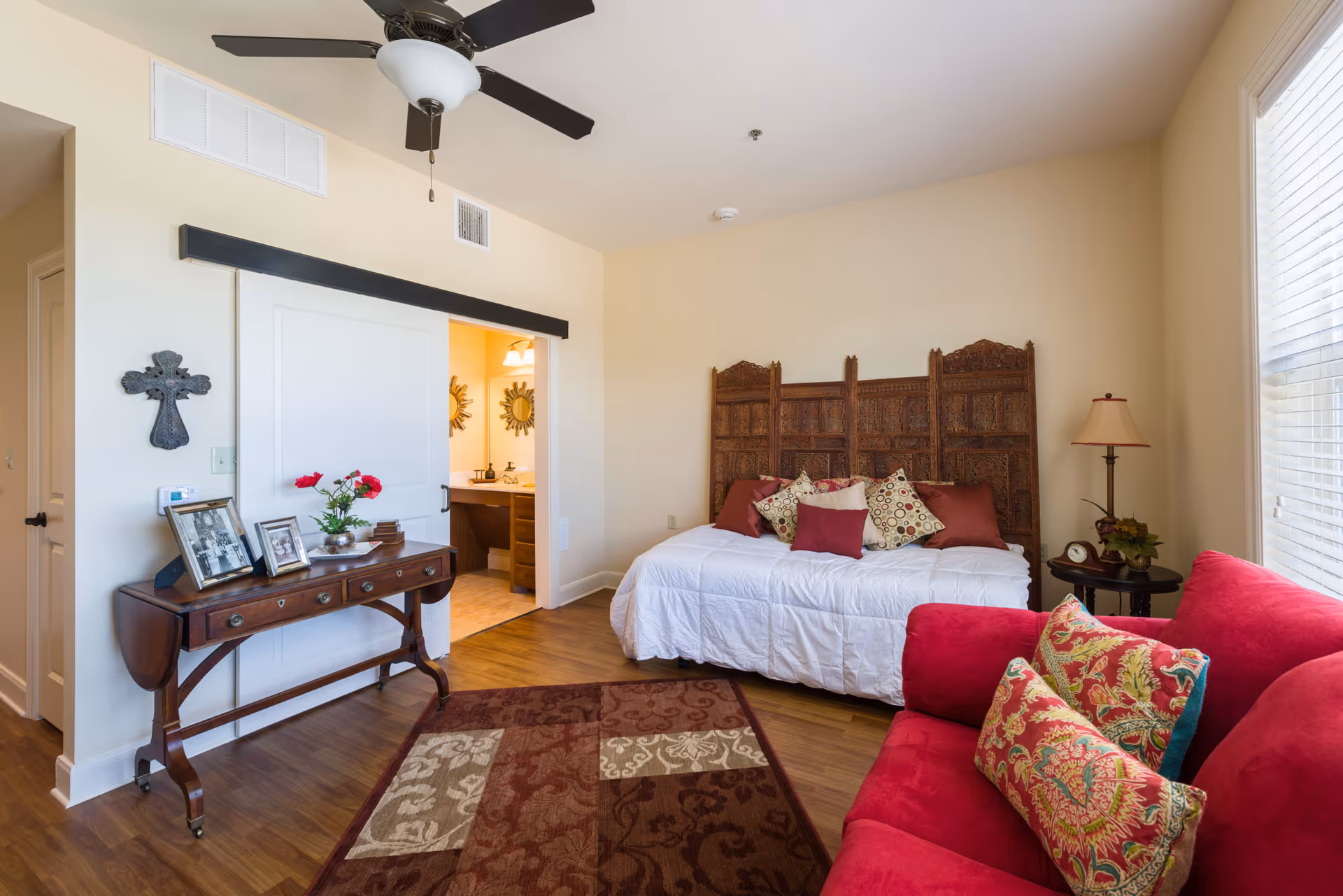 Bright furnished living area with a daybed, red sofa, wooden console table, patterned rug and decorative room divider under a ceiling fan.